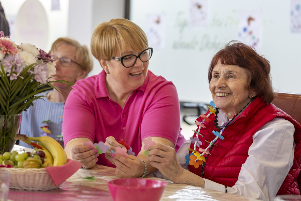 Seniorin und Mitarbeiterin sitzen am Tisch und lachen im Gespräch – häusliche Pflege in vertrauter Umgebung