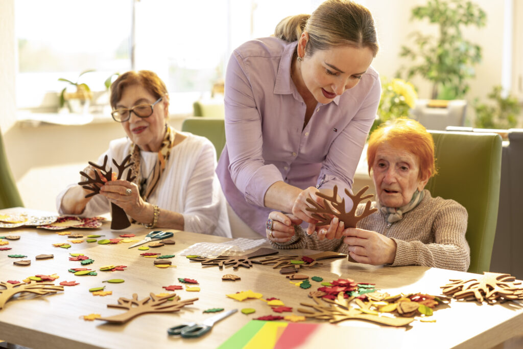 Seniorin und Mitarbeiterin im Gespräch am Tisch; Notizen und Getränk liegen bereit – Start der häuslichen Pflege.