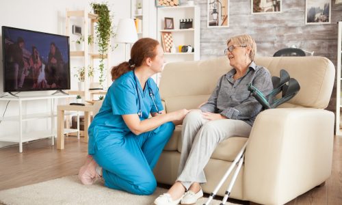 Pflegekraft kniet vor Seniorin auf dem Sofa und hält ihre Hand – einfühlsame ambulante Betreuung zu Hause.